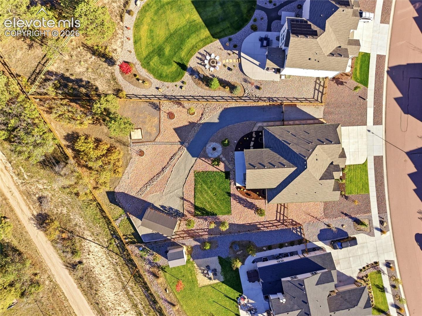 Aerial perspective featuring a multi-pitched roofline, landscaped grounds with designated lawn areas, perimeter fencing, gravel pathways, and a detached storage structure