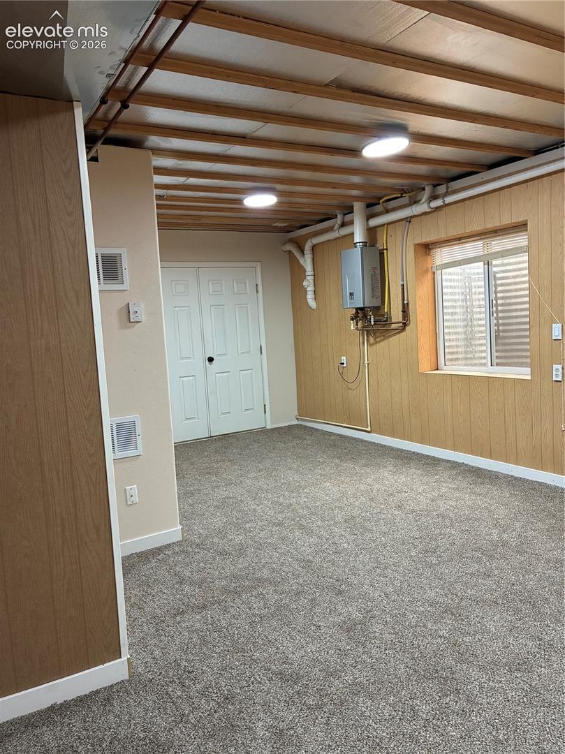 Finished interior space featuring exposed joist ceiling, wall-mounted tankless water heater, wood-finish wall paneling, carpeted flooring, and double-door entry