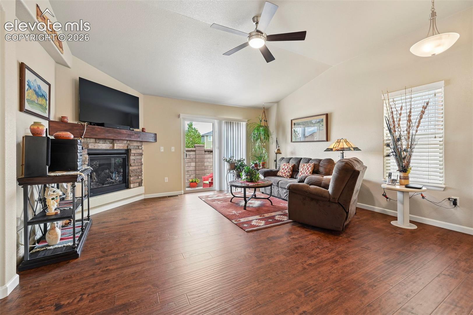 Living room featuring vaulted ceilings, vinyl flooring, a stone fireplace with a substantial wood mantel, a ceiling fan, and sliding glass door to private patio