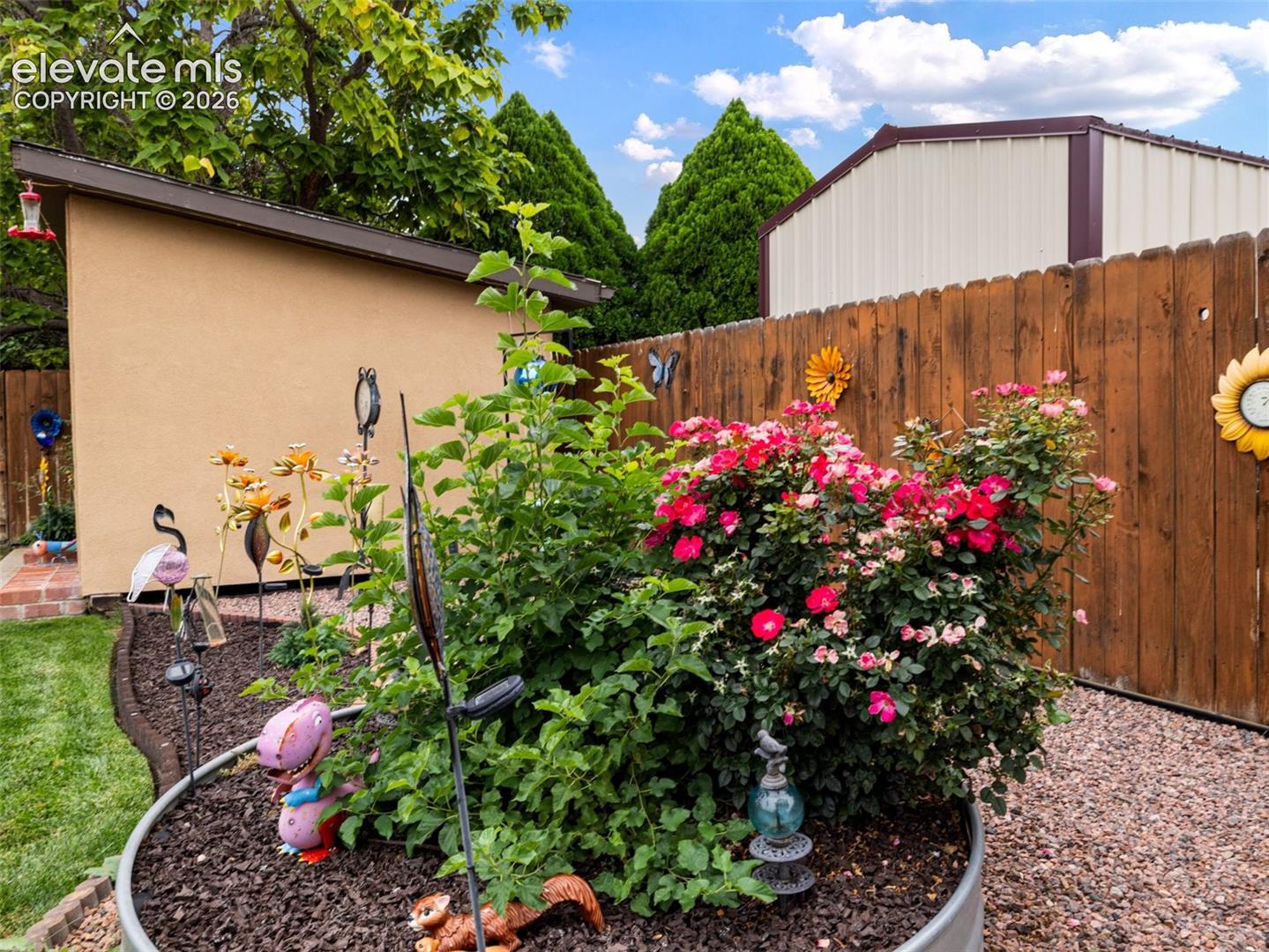 Stucco exterior structure, wood privacy fencing, mature green foliage, vibrant pink and red flowering shrubs, and a gravel ground cover