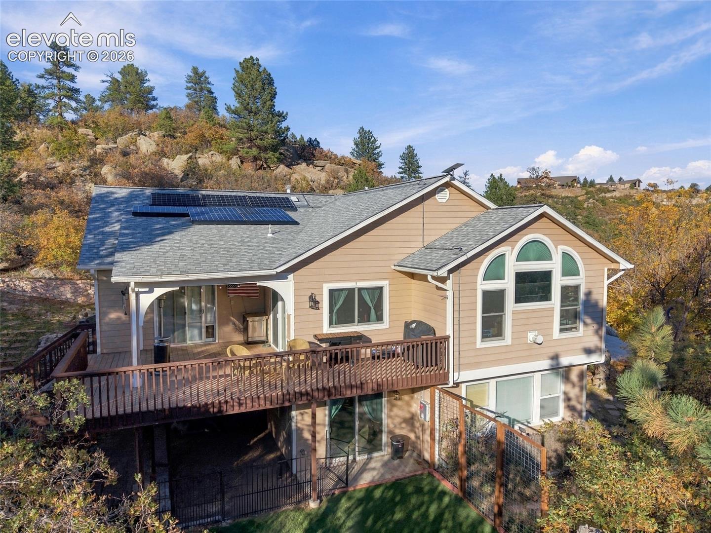 Multi-level exterior featuring a raised wooden deck with railings, solar panels, arched windows, and a fenced lower-level patio area