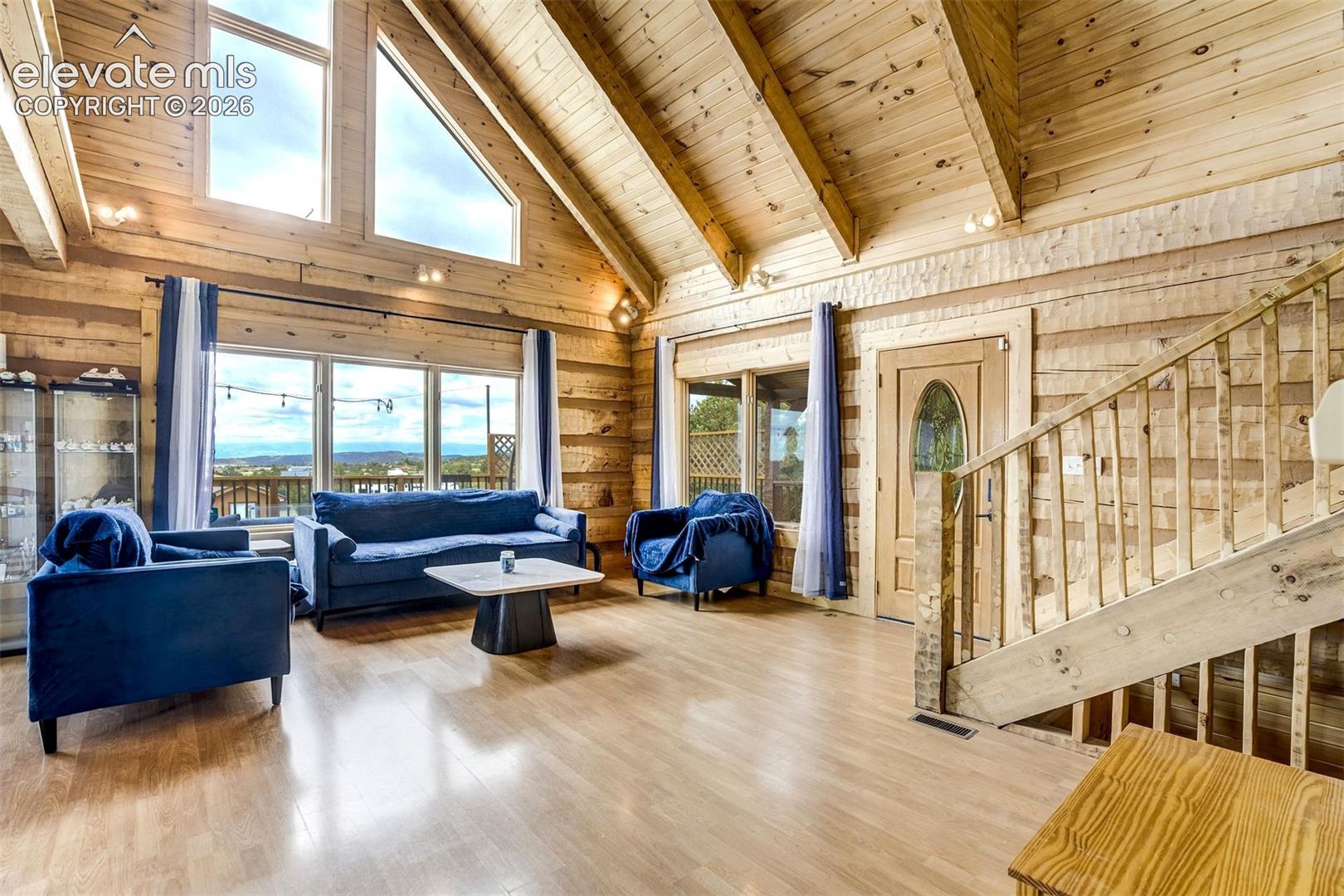 Spacious living area featuring expansive windows, wood-finish flooring, exposed beam ceilings, and a wood-paneled accent wall