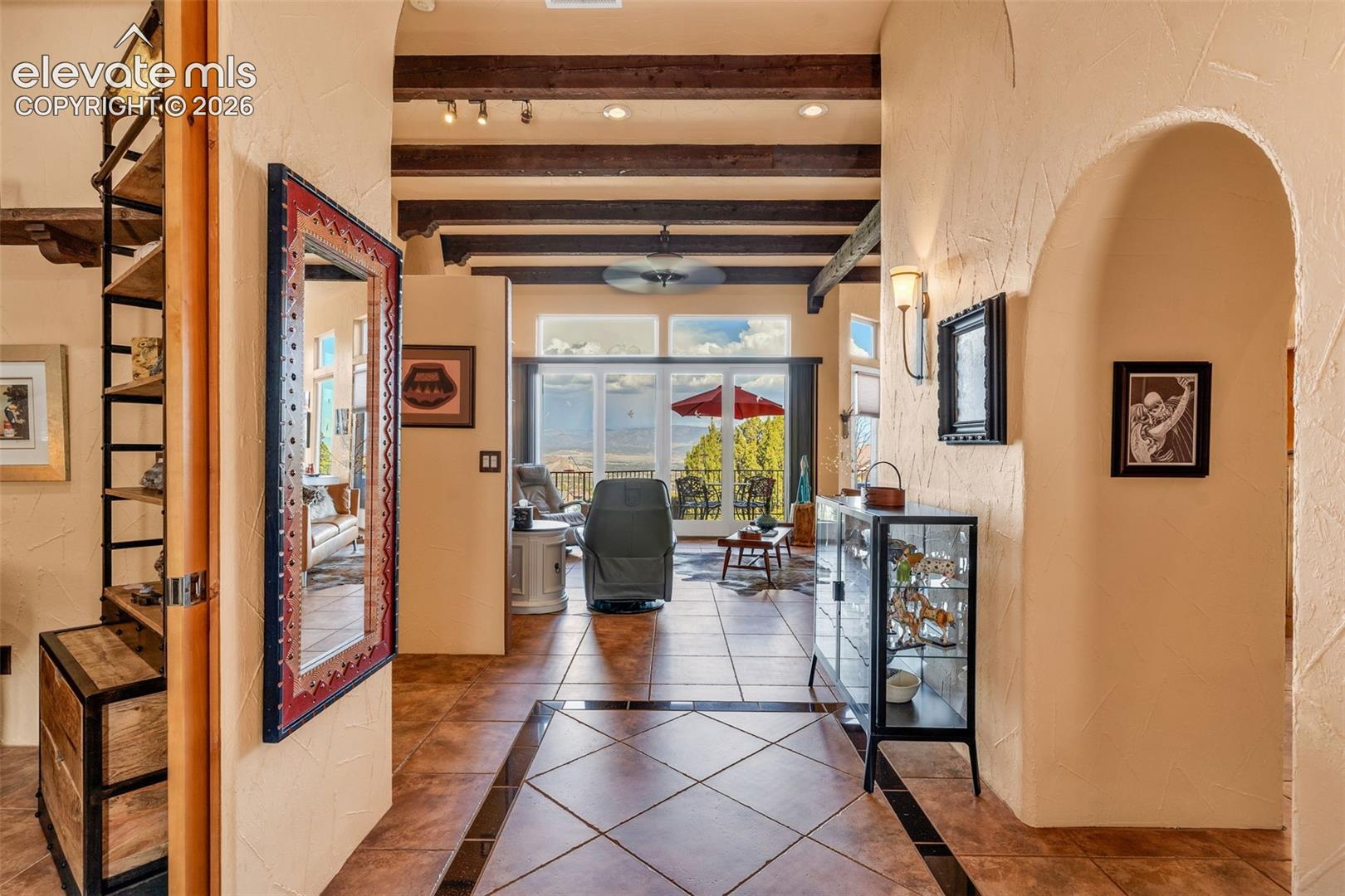 Interior space featuring exposed wooden ceiling beams, arched doorways, textured plaster walls, ceramic tile flooring with decorative inlay, and expansive windows