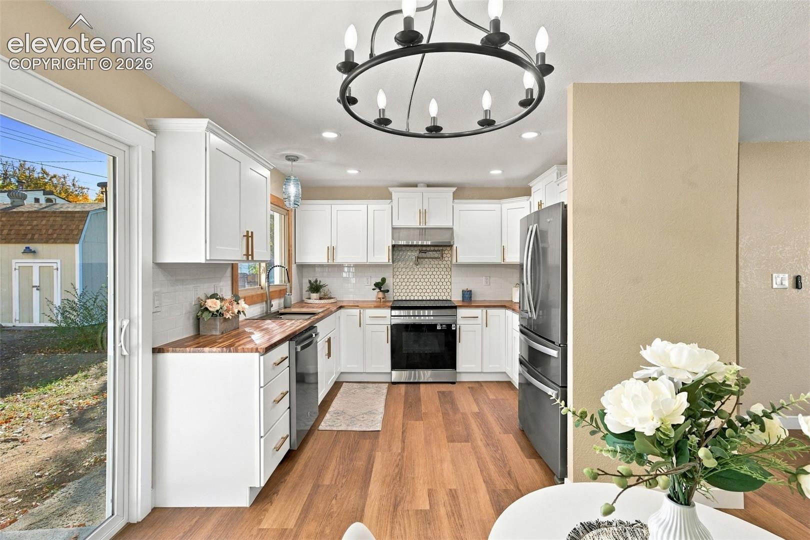Kitchen featuring white cabinetry with brushed-gold hardware, wood-finish countertops, stainless steel appliances, a hexagonal tile backsplash, and a circular wagon-wheel style chandelier