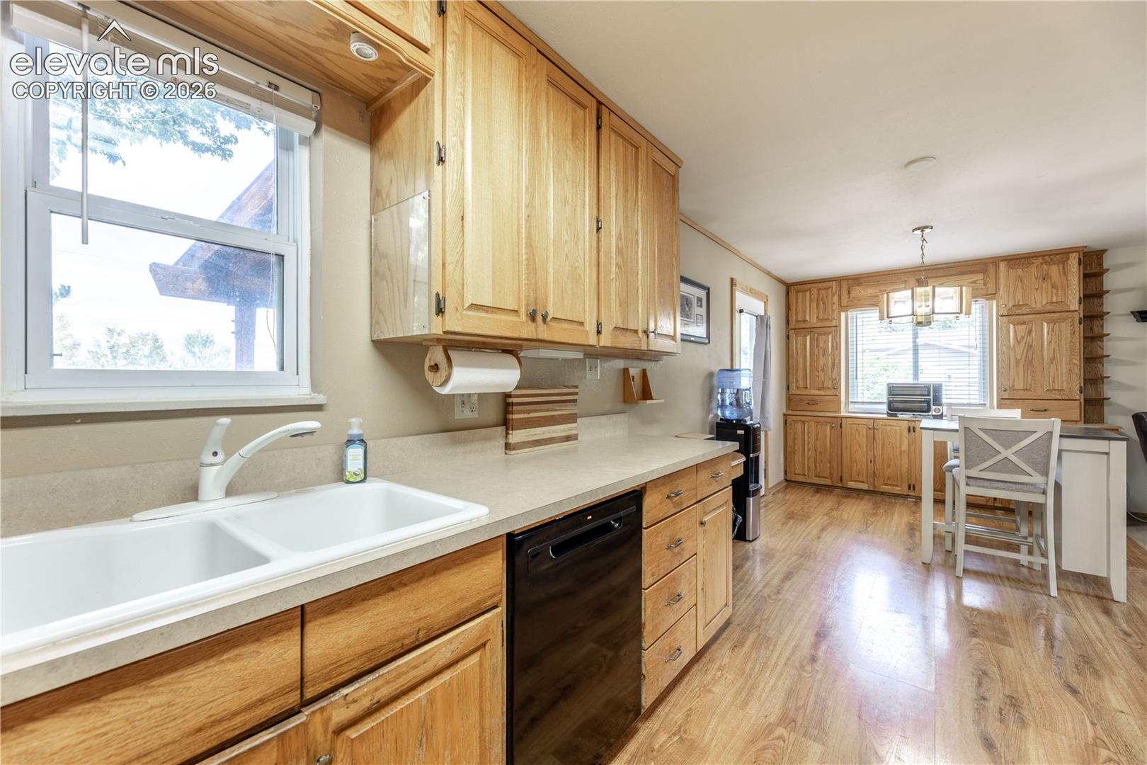 Kitchen featuring light wood-finish cabinetry, a white dual-basin sink with a high-arch faucet, a black dishwasher, extensive built-in cabinetry, and wood-finish flooring
