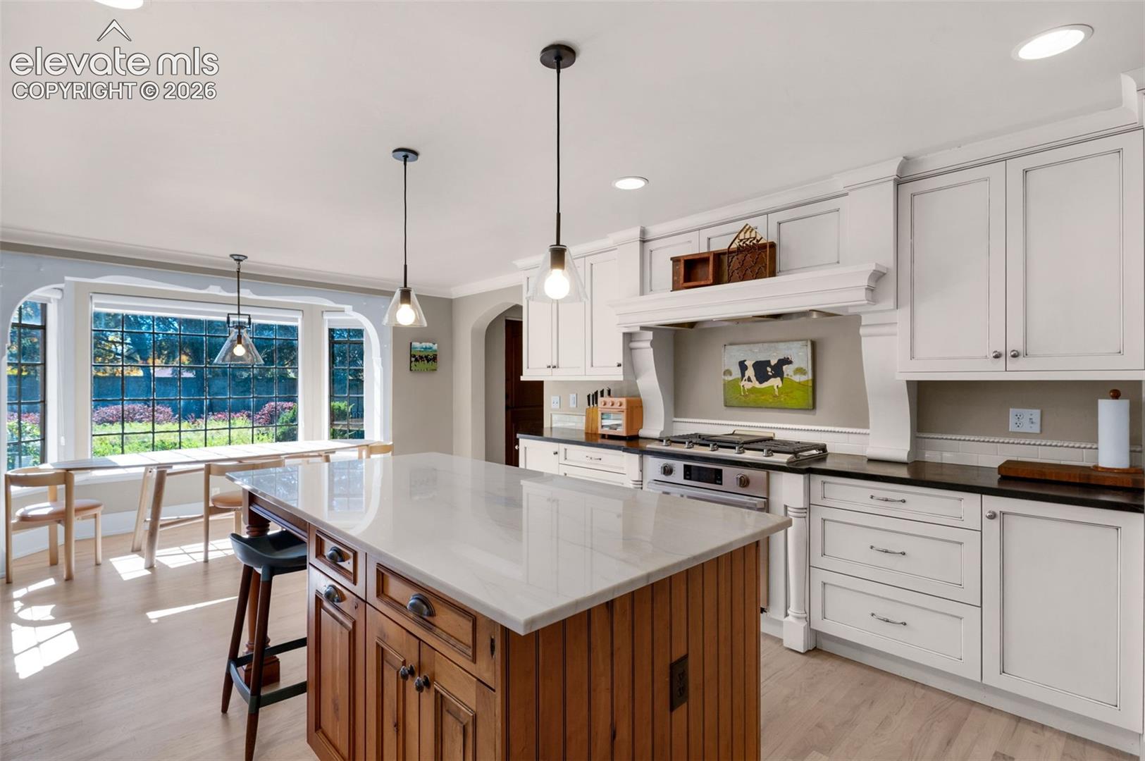 Kitchen featuring a wood-finish island with a light-toned countertop, custom white cabinetry, built-in range hood, pendant lighting, and large bay windows