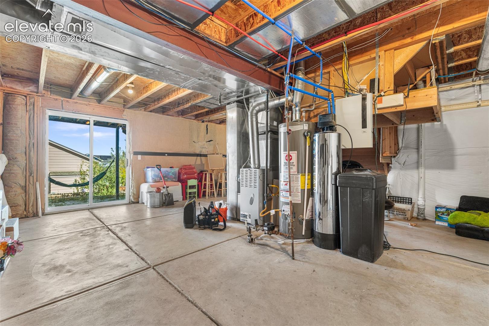 Unfinished basement area featuring a concrete floor, exposed ceiling joists, water heating system, water filtration unit, and a sliding glass door