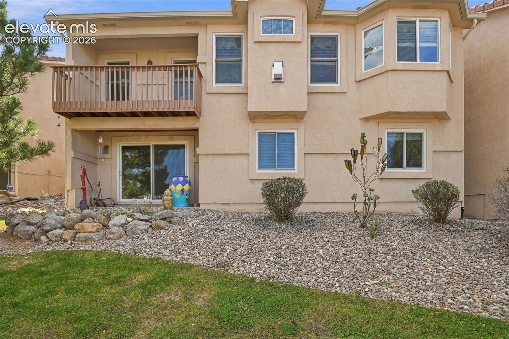 Stucco exterior featuring an upper-level balcony with wood railing, sliding glass doors, and xeriscaped grounds with a stone retaining wall
