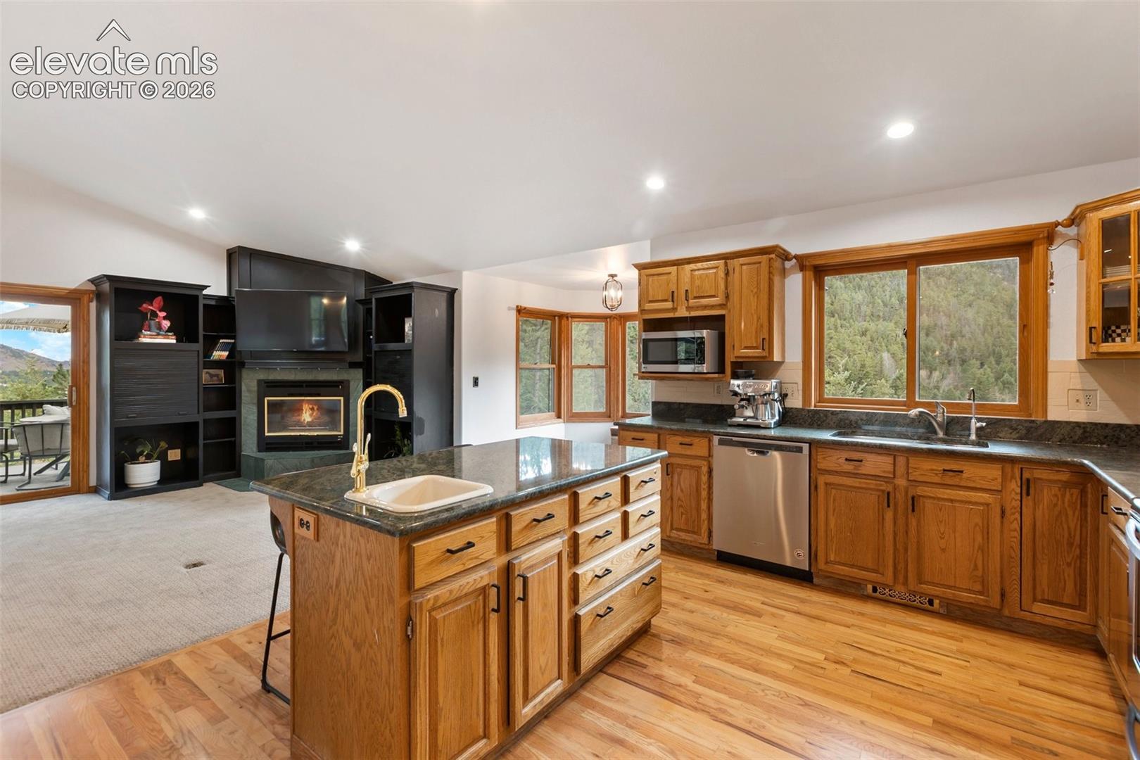 Kitchen featuring wood cabinetry, stone countertops, central island with inset sink, stainless steel dishwasher, and built-in fireplace with integrated shelving