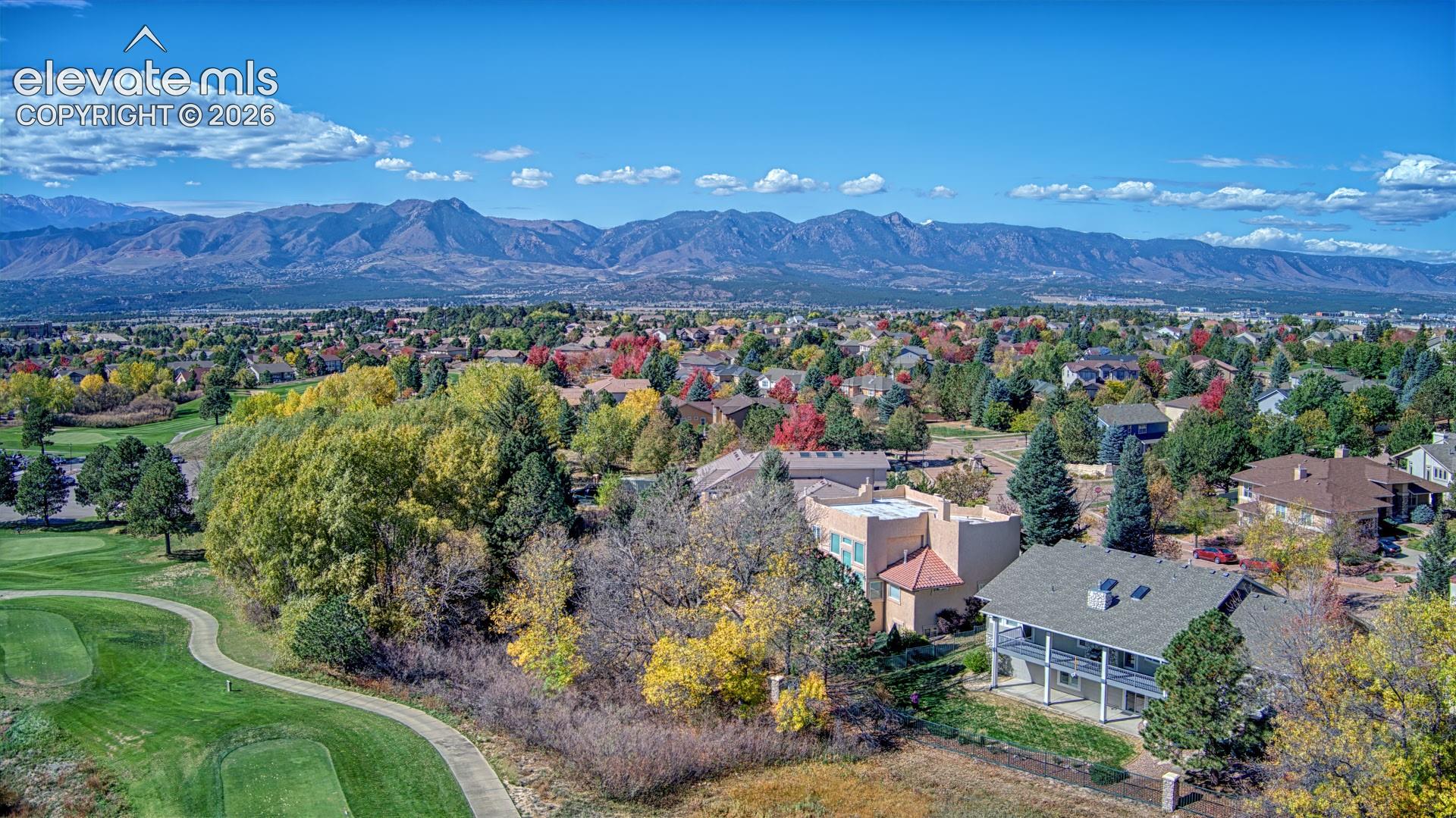 Aerial perspective featuring a multi-story residence with a covered rear patio and balcony, situated on a lot bordering a golf course with mature trees and a prominent mountain range backdrop