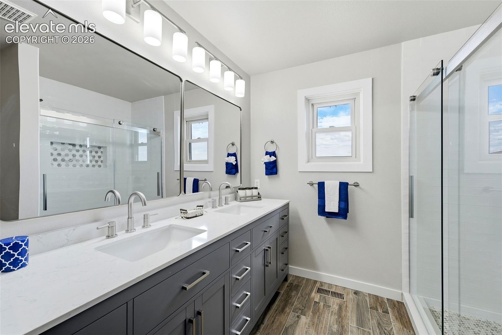Dual-sink vanity with a light countertop, wood-finish flooring, and a glass-enclosed shower featuring a tiled niche