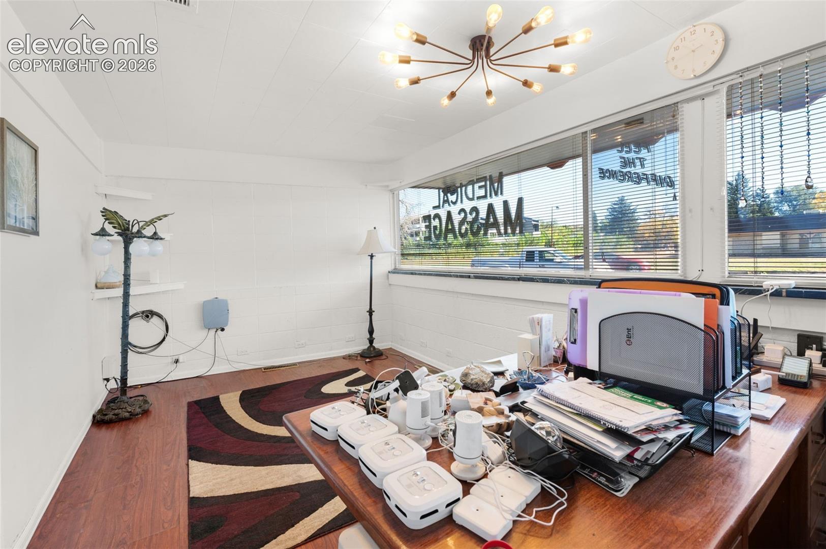 Spacious room featuring wood-finish flooring, white masonry walls, large picture windows with blinds, and a multi-arm sputnik-style ceiling light fixture