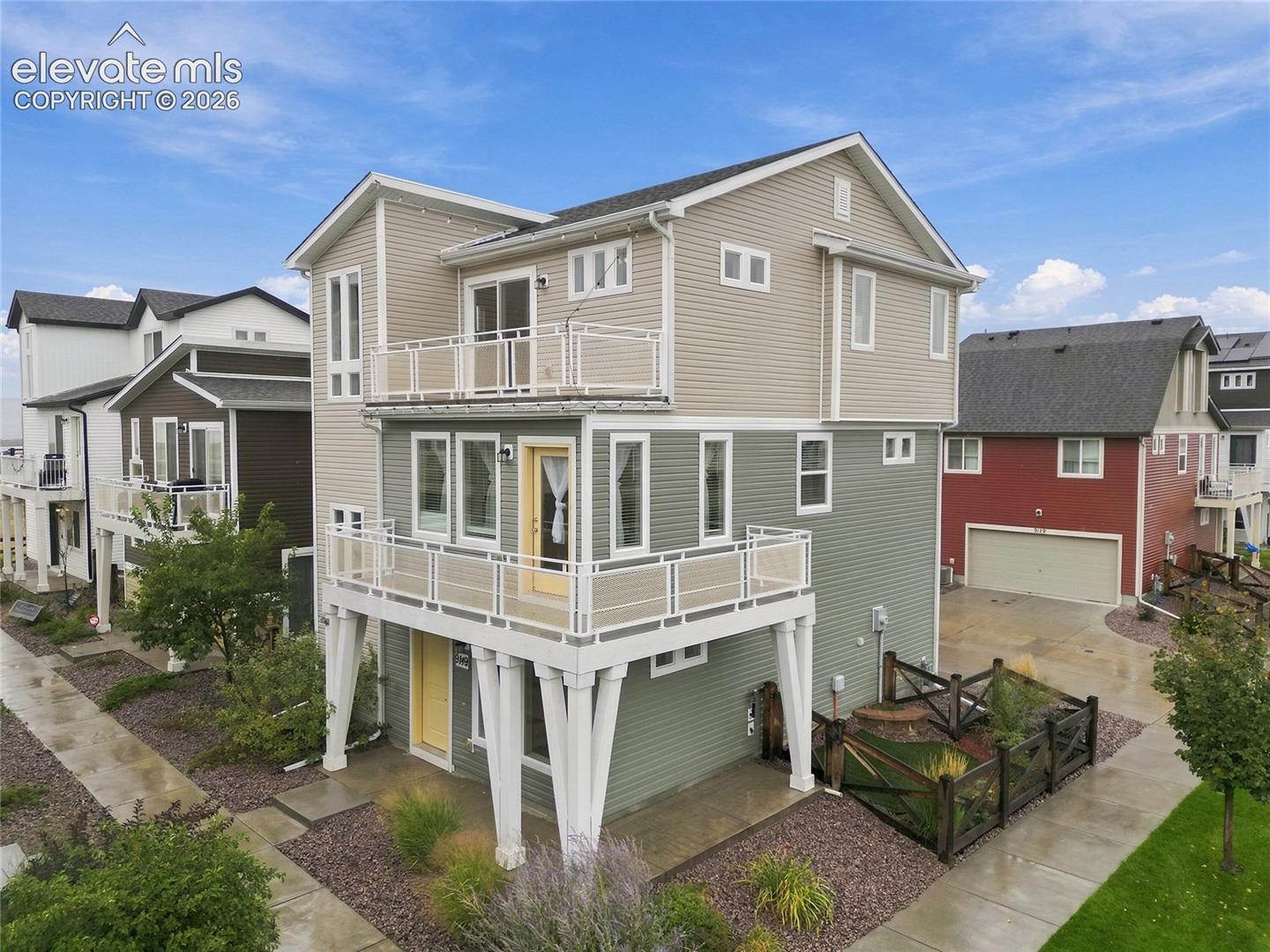 Multi-level residence with two balconies featuring white railings, an elevated main entrance with a yellow door, and a dedicated garden area with fencing