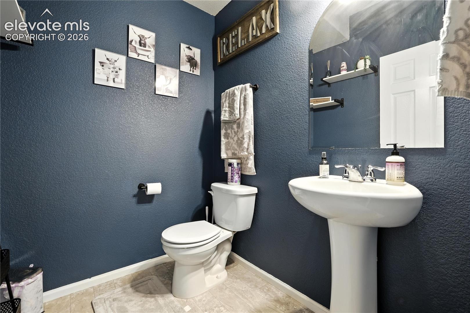 Pedestal sink with chrome fixtures, an arched mirror, and wall-mounted shelving