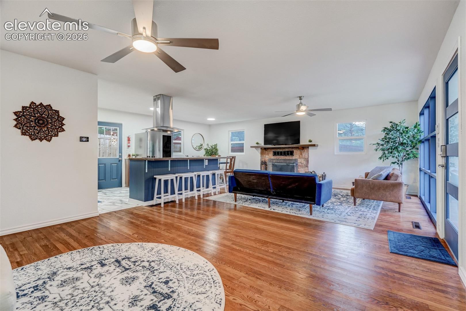 Wood-finish flooring throughout the main area, stone fireplace, kitchen island with seating, stainless steel range hood, and ceiling fans