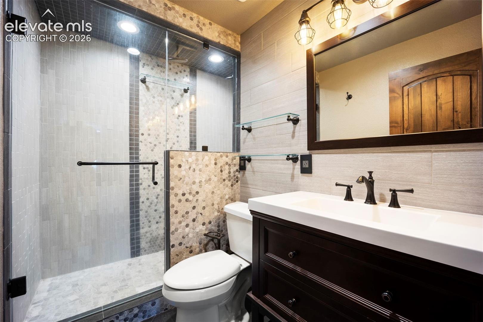 Bathroom featuring a glass-enclosed shower with tile surround, a dark wood vanity with white countertop, a framed mirror, and industrial-style vanity lighting