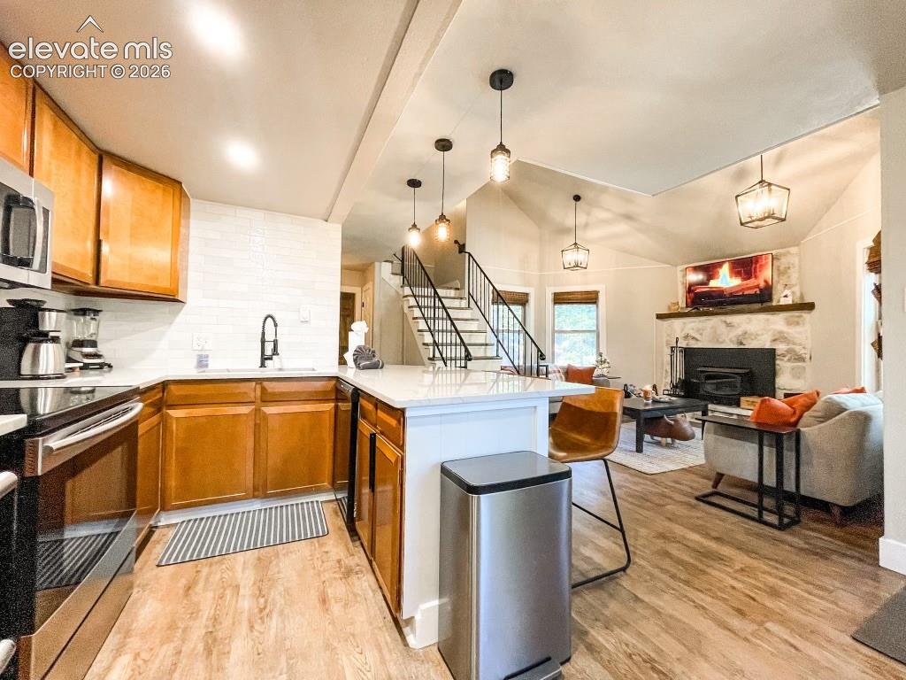 Open-concept kitchen and living area featuring wood-finish flooring, a kitchen island with seating, stainless steel appliances, and a living room with a vaulted ceiling and a stone fireplace
