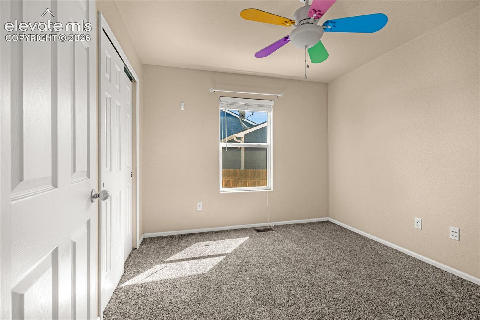 Neutral wall paint, gray carpet, paneled closet door, window with blinds, and a ceiling fan