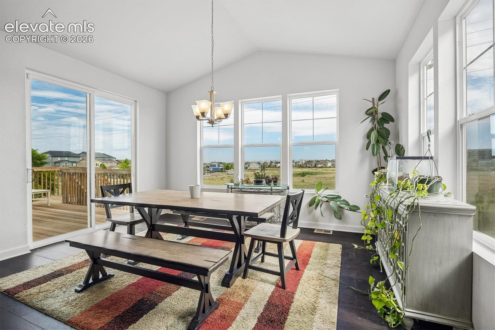 Vaulted ceiling dining area featuring rich wood-finish flooring, a contemporary chandelier, multiple large windows, and a sliding glass door opening to a private deck