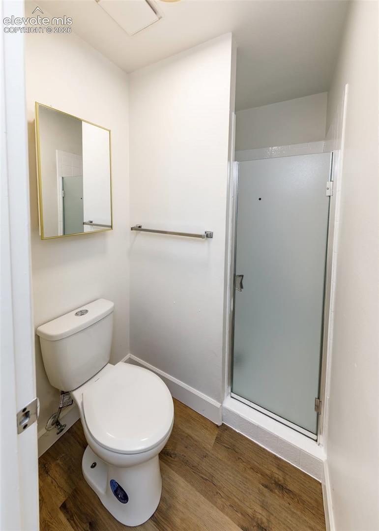 Bathroom featuring wood-finish flooring, a wall-mounted rectangular mirror with a gold-tone frame, a ceramic toilet, and a shower enclosure with a frosted glass door