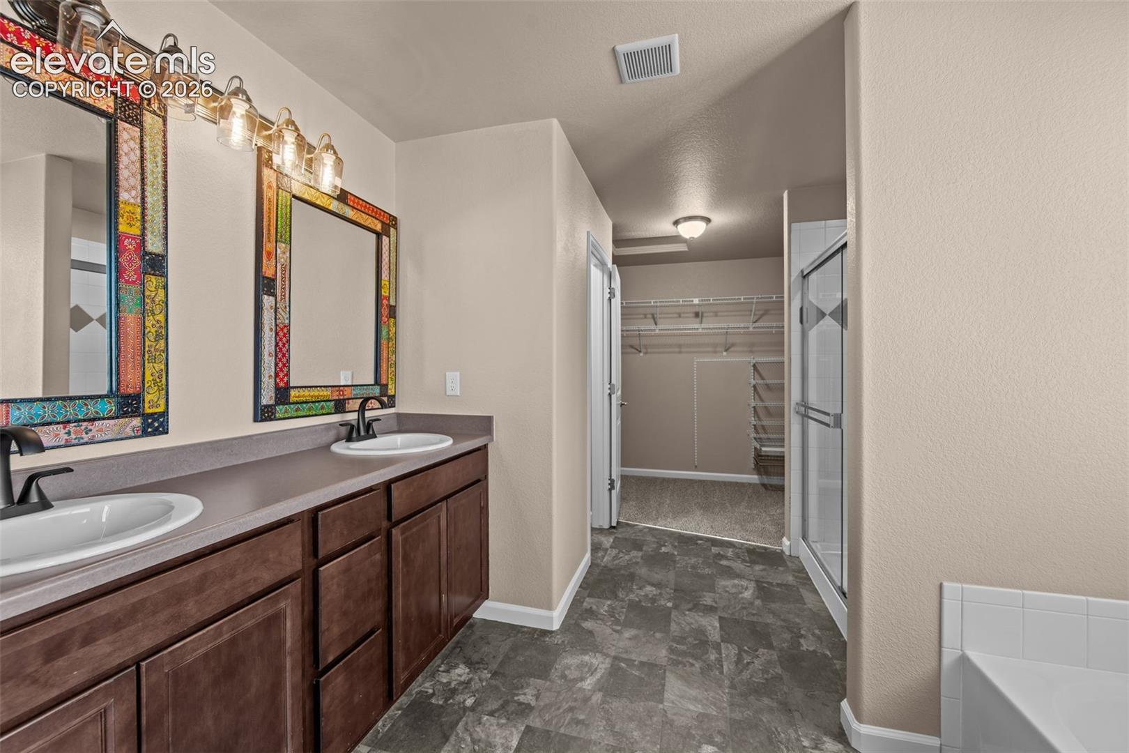 Dual-sink vanity with dark wood cabinetry, solid-surface countertop, matte black fixtures, tile flooring, and integrated walk-in storage