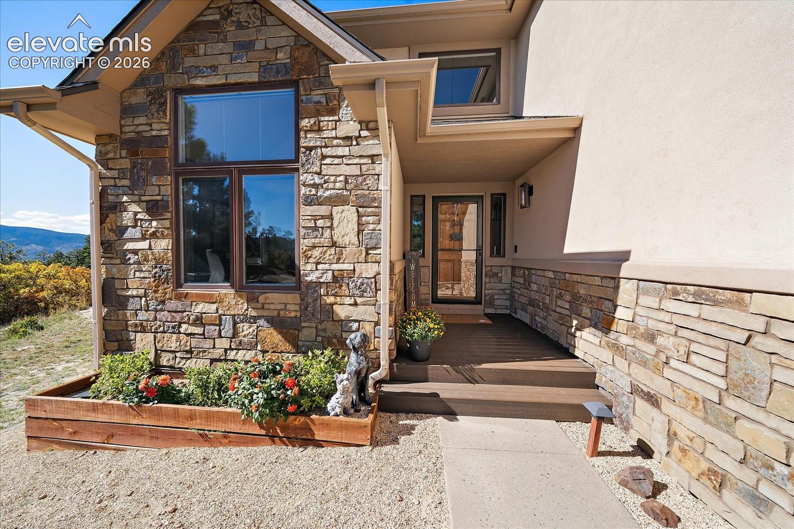 Exterior facade featuring multi-textured stone masonry, stucco siding, a covered entryway with wood-finish decking, and large-format glass window panes