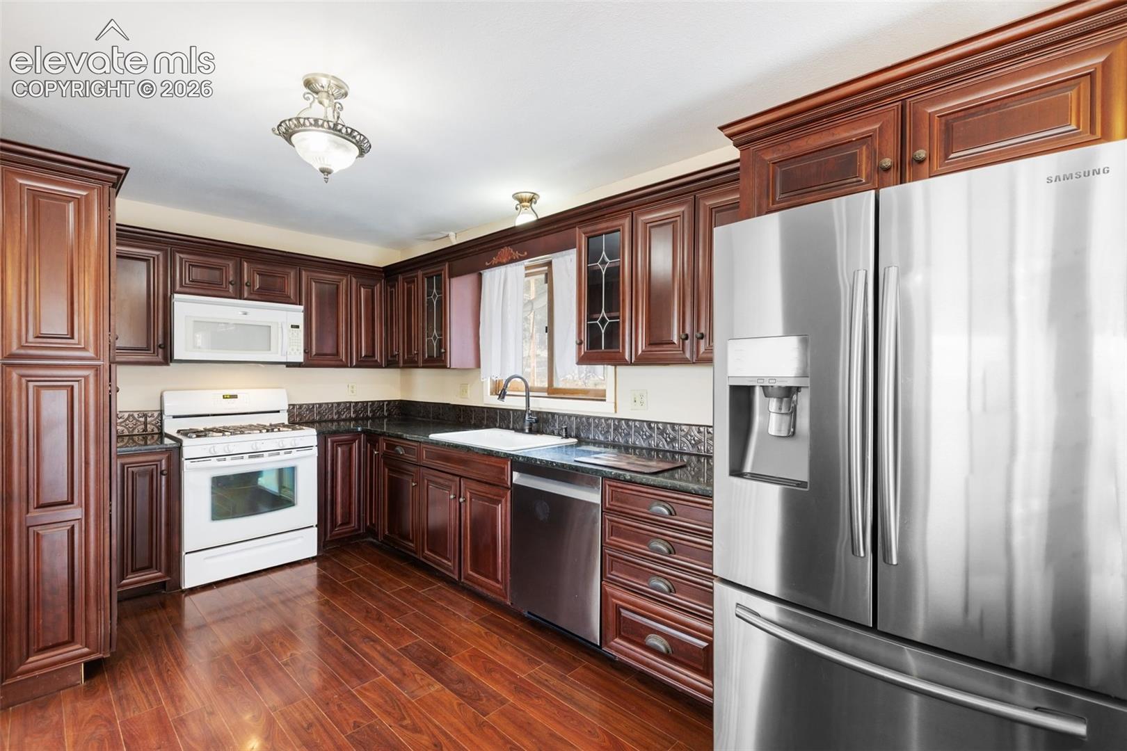 Rich wood cabinetry with crown molding, stainless steel appliances, dark stone countertops, wood-finish flooring, and an undermount sink