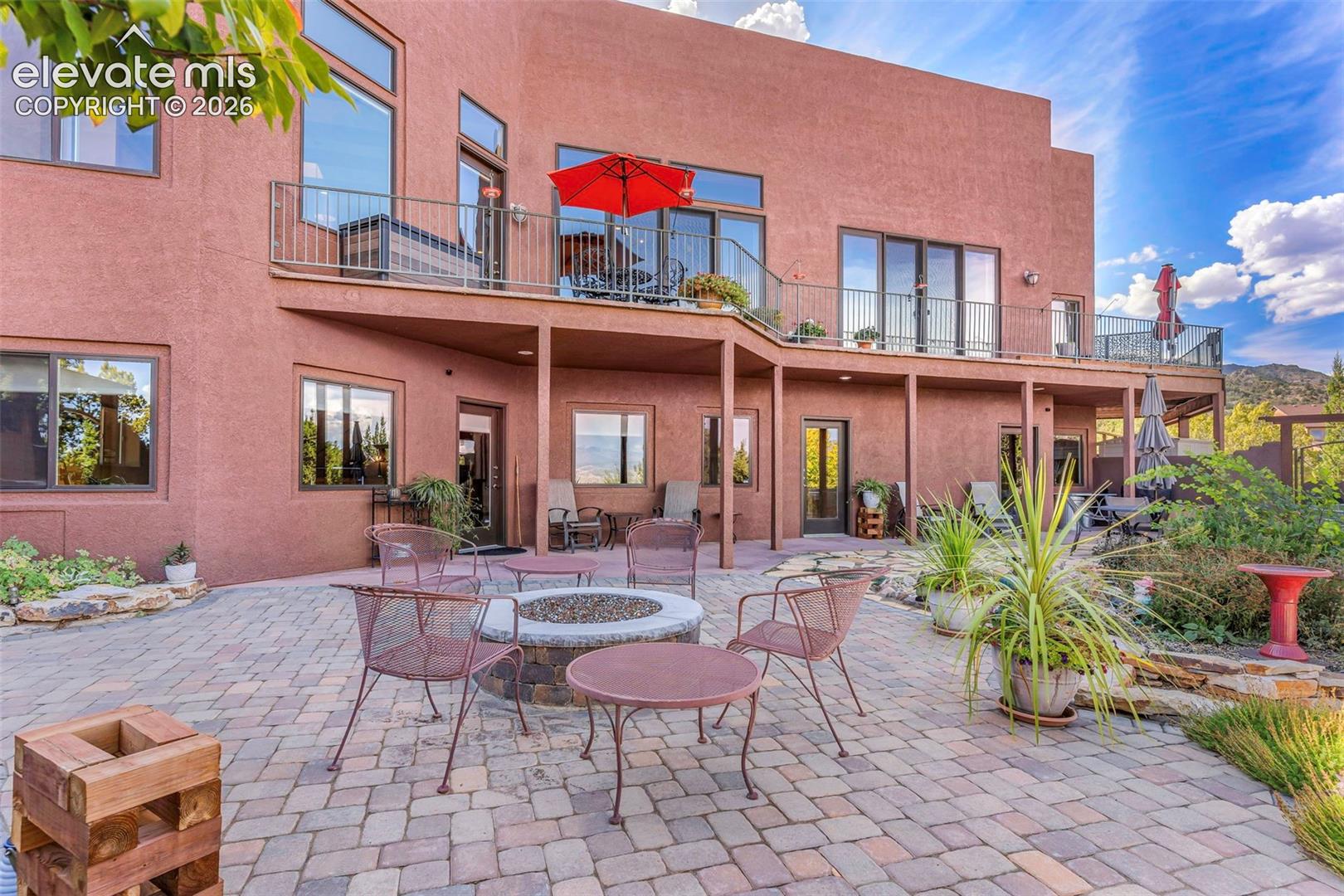 Stucco exterior featuring a wraparound elevated deck with metal railings and a ground-level paver patio with a built-in stone fire pit