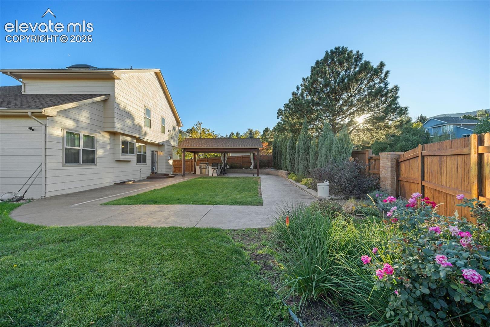 Backyard featuring a concrete patio, covered outdoor pavilion, manicured lawn, perimeter wood fencing, and established perennial landscaping