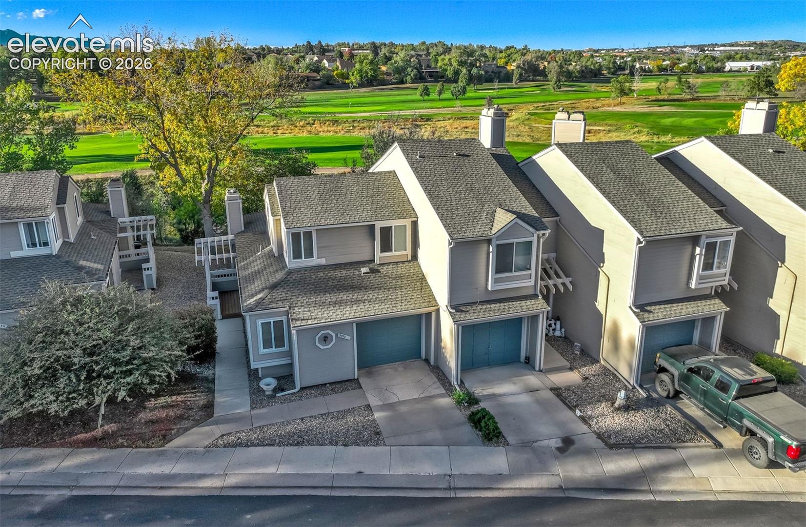 Multi-level townhome exterior featuring shingled rooflines, attached garages, light-toned siding, and exterior pergolas, situated against a backdrop of open green space