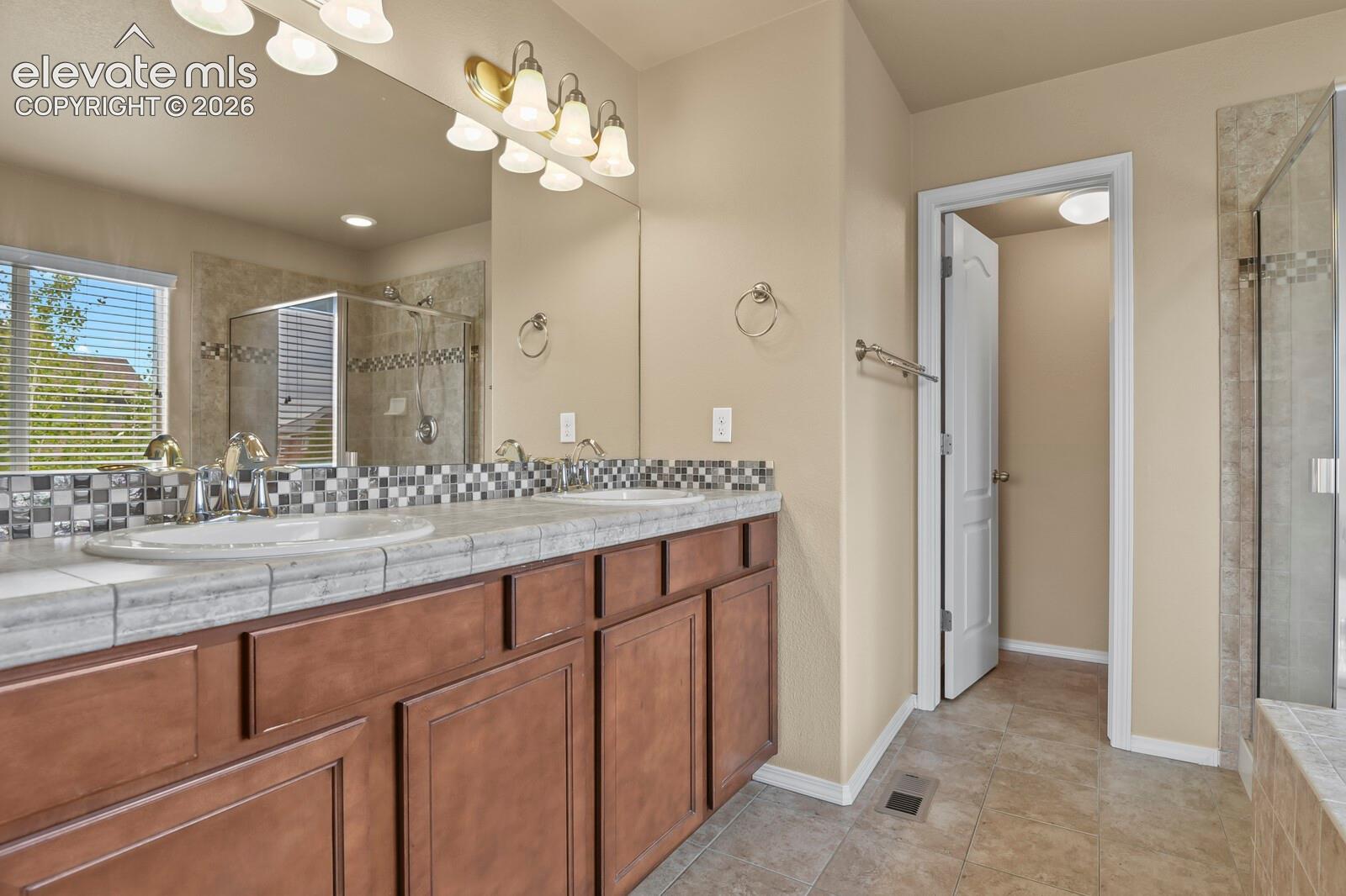 Dual vanity with wood-finish cabinetry, tile countertop, and mosaic tile backsplash