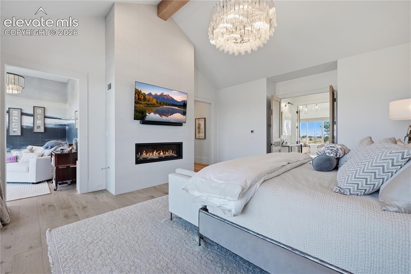 Vaulted ceilings with exposed timber beams, linear gas fireplace with full-height surround, wood-finish flooring, and multi-tier crystal chandelier