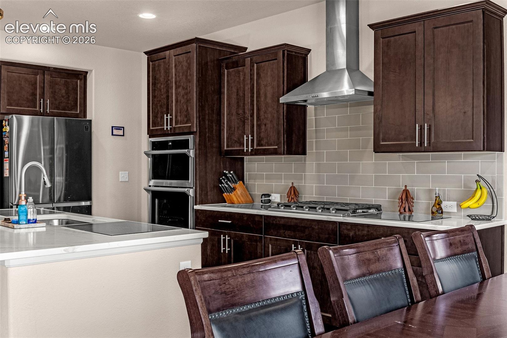 Kitchen featuring dark wood cabinetry, stainless steel appliances, a stone-finish countertop, subway tile backsplash, and a wall-mounted stainless steel vent hood