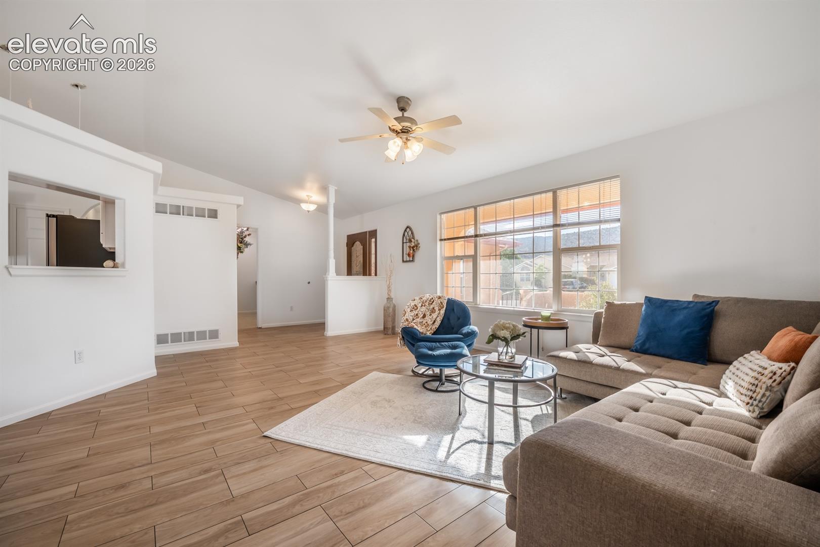 Living area featuring wood-finish flooring, vaulted ceilings, large multi-pane windows, and a ceiling fan with integrated lighting