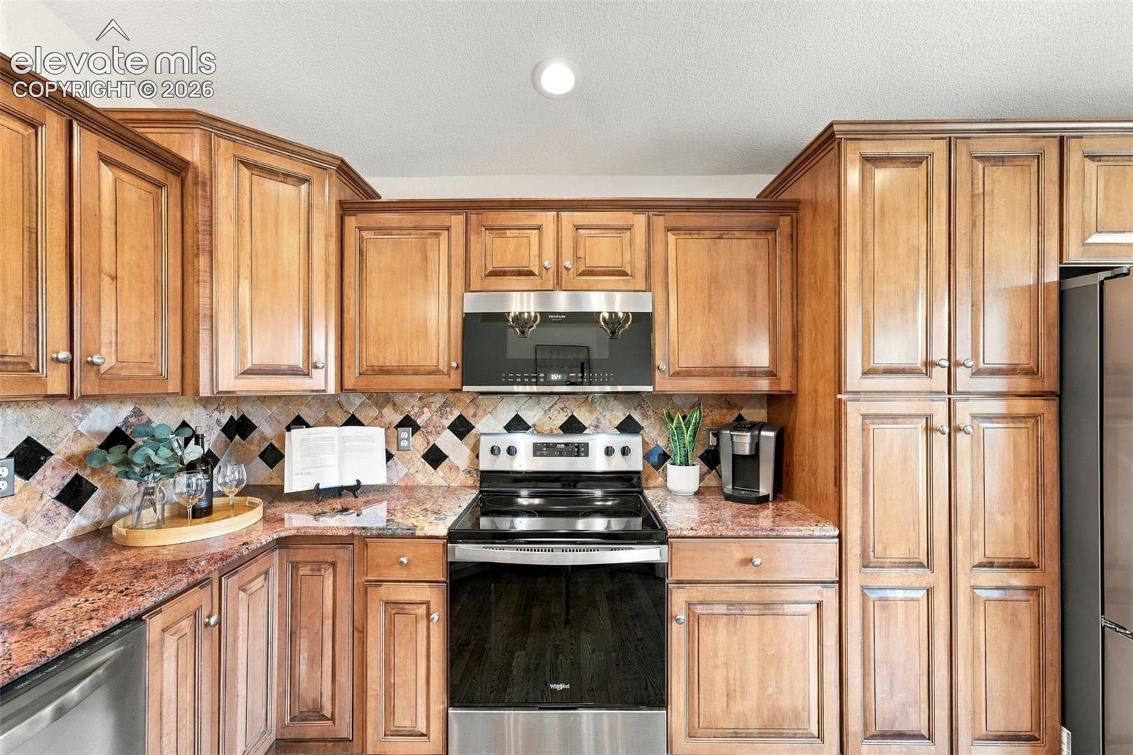Raised-panel wood cabinetry, natural stone countertops, geometric tile backsplash, and stainless steel appliances