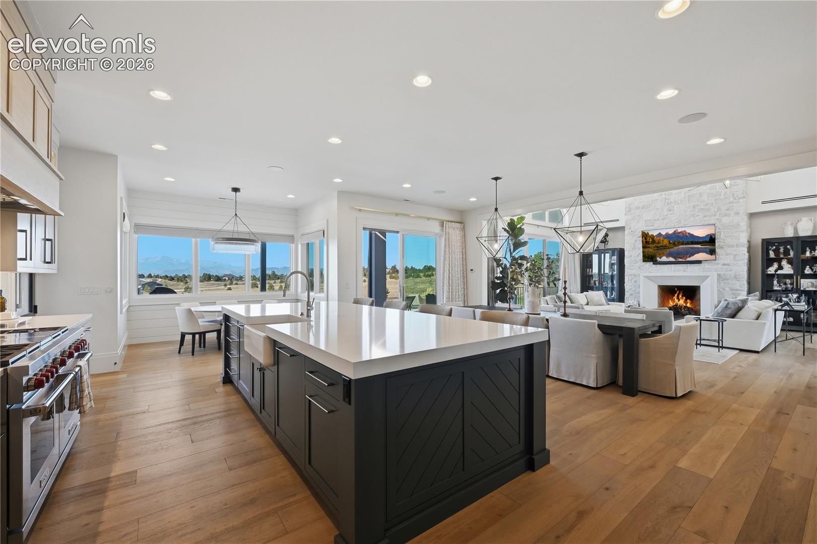 Gourmet kitchen featuring a large central island with quartz countertops, custom cabinetry with herringbone detailing, wood-finish flooring, recessed lighting, and a stone-clad fireplace