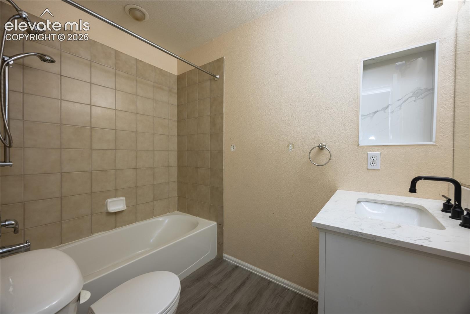 Full bathroom featuring a soaking tub with tile surround and overhead shower, wood-finish flooring, single-sink vanity with white countertop and matte black faucet, toilet, and recessed lighting