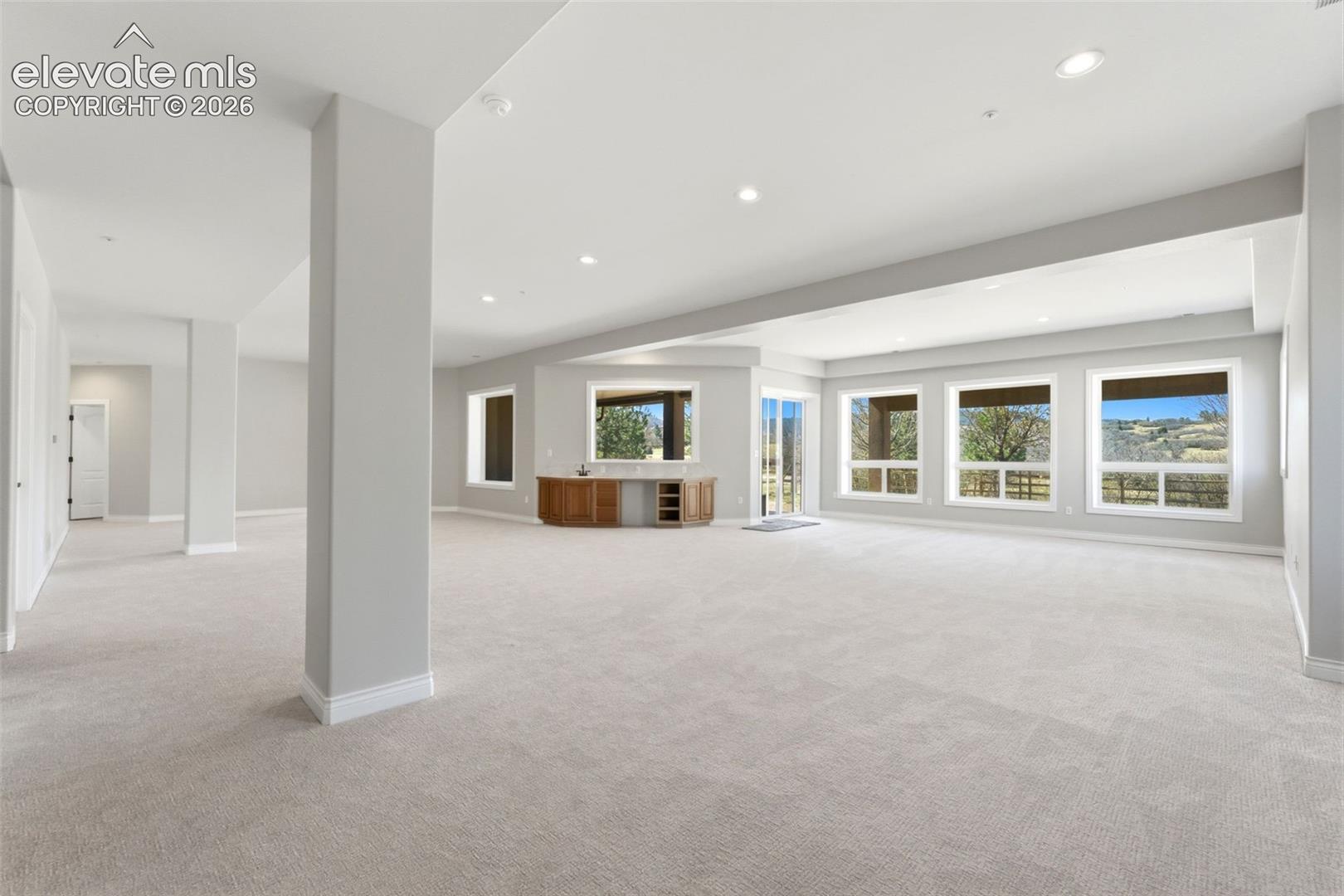Expansive open-concept room featuring light-toned carpeting, recessed lighting, a built-in wood-finish wet bar, and multiple large windows with exterior scenery