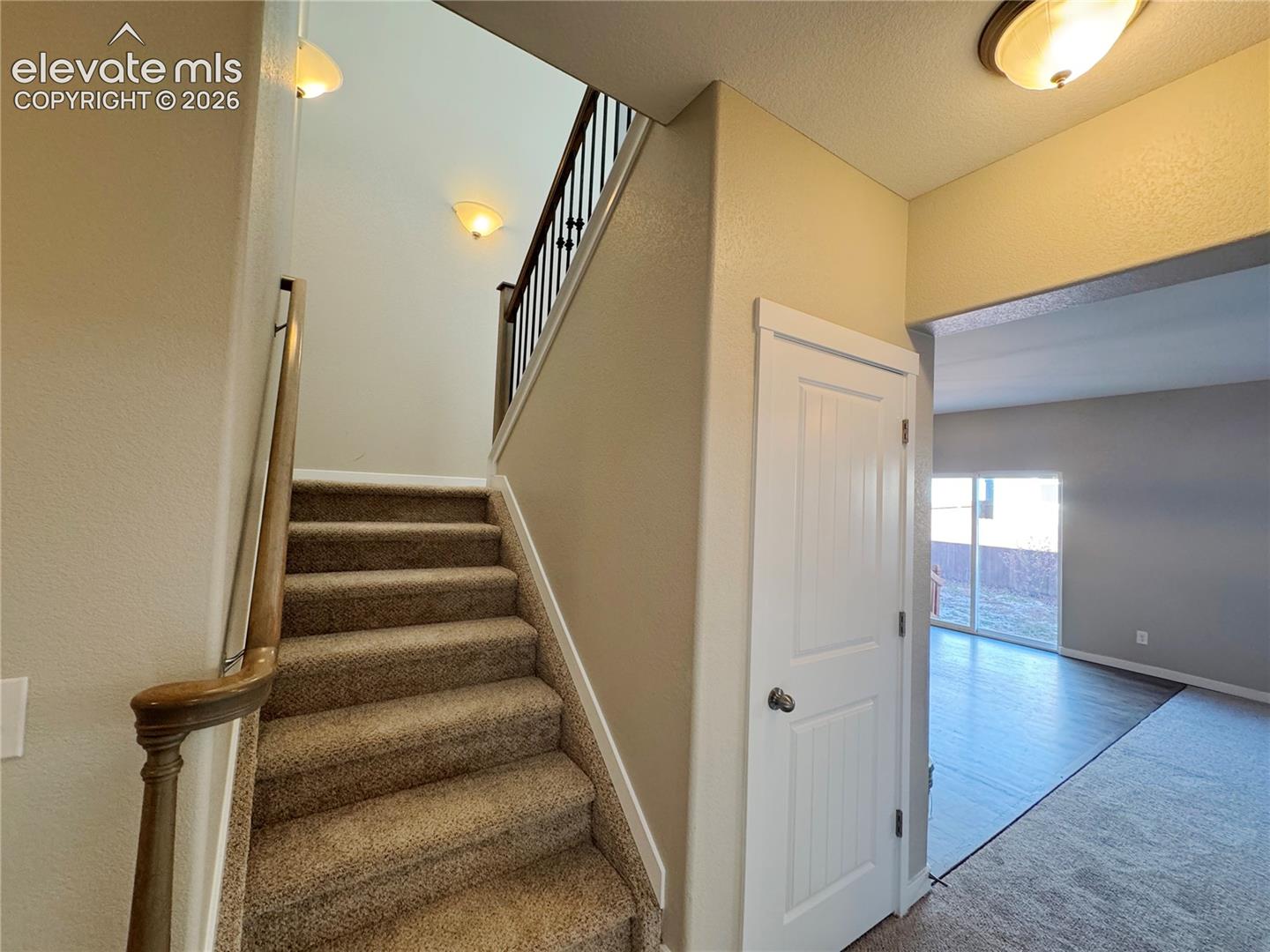 Staircase featuring a wood handrail with metal balusters, carpeted treads, a solid-core interior door, and flush-mount ceiling lighting