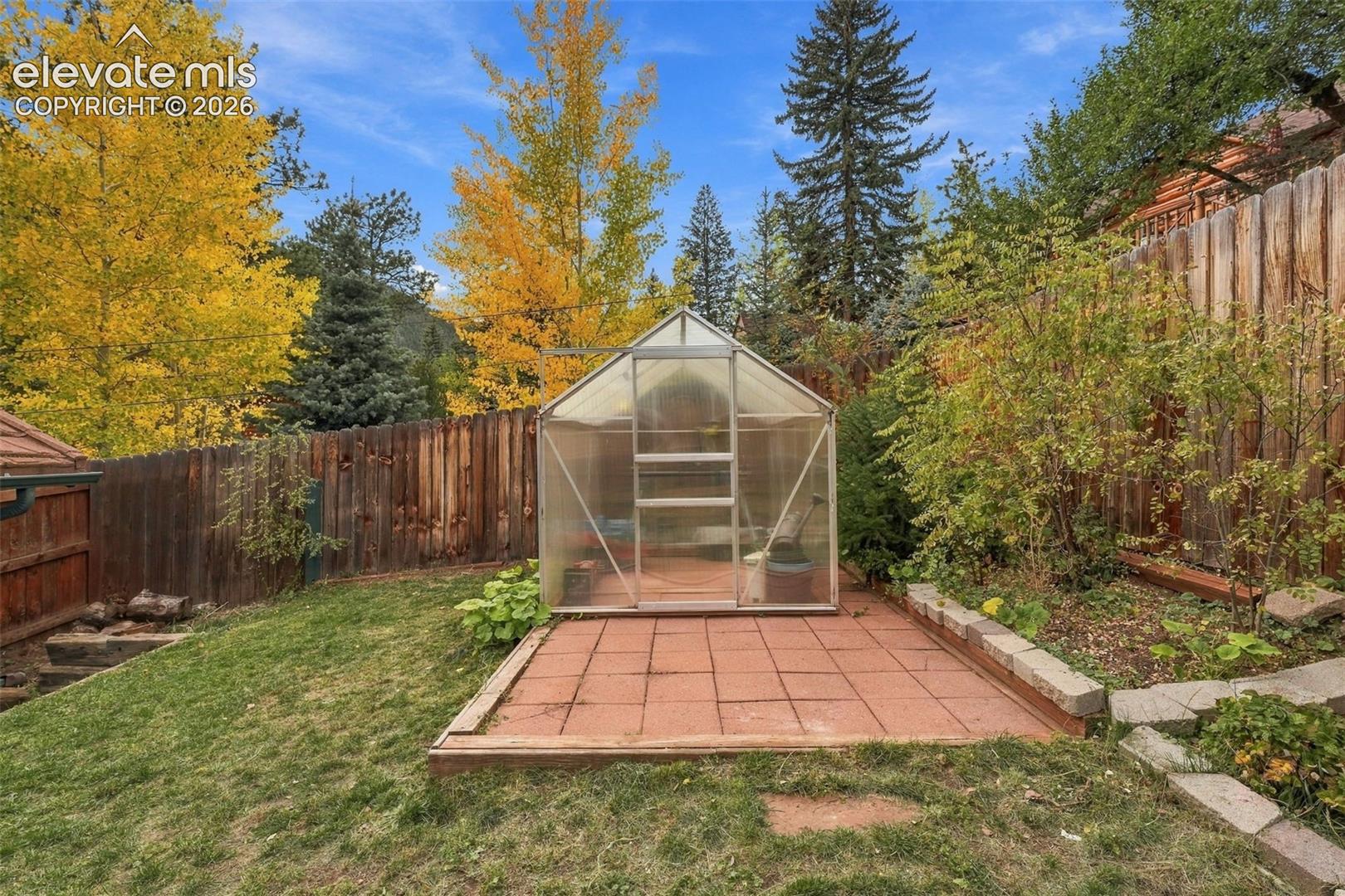 Freestanding polycarbonate greenhouse situated on a red masonry paver patio, enclosed by a wood privacy fence and mature landscaping