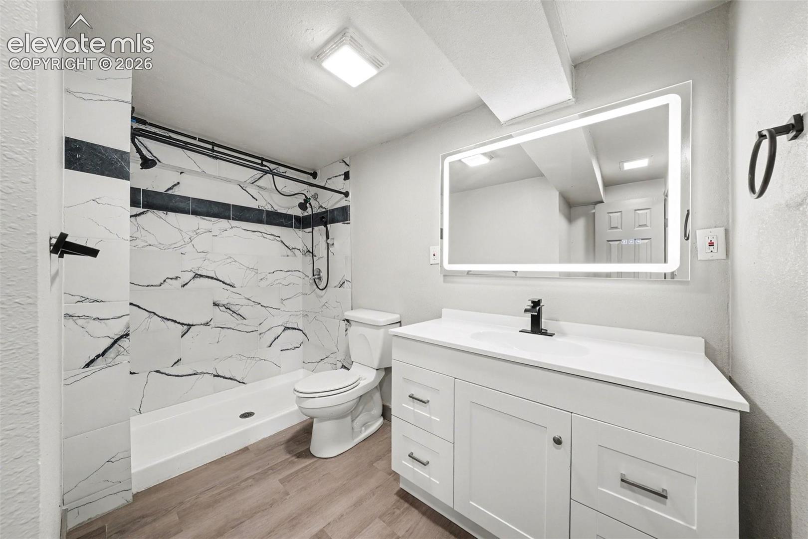 White vanity with integrated basin and matte black faucet, illuminated vanity mirror, wood-finish flooring, and walk-in shower featuring large-format patterned wall tile with dark accent inlay