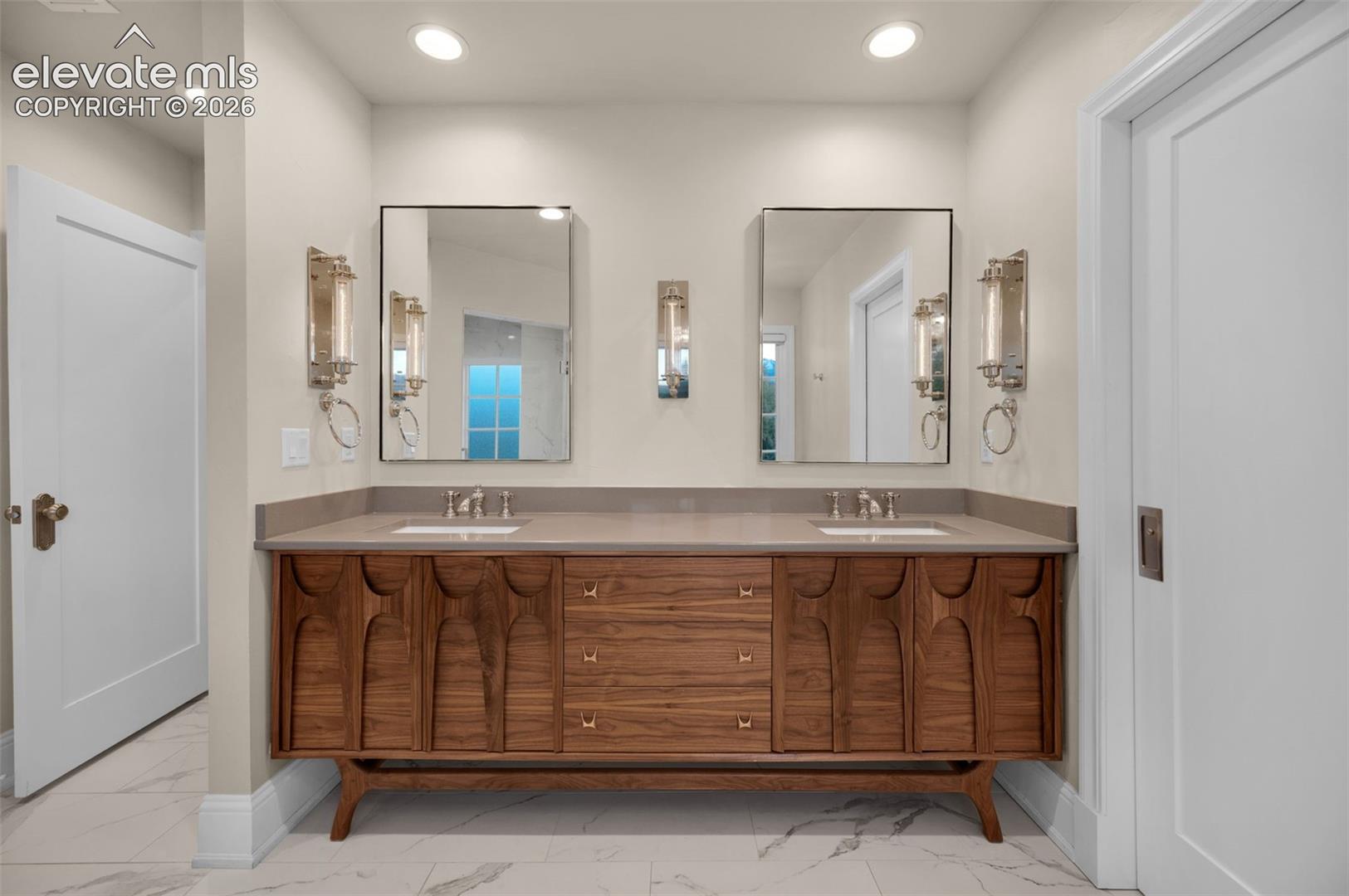 Mid-century modern dual-sink vanity featuring warm-toned wood cabinetry with decorative carved detailing, solid-surface countertop, rectangular undermount sinks, and metallic wall-mounted light fixtures