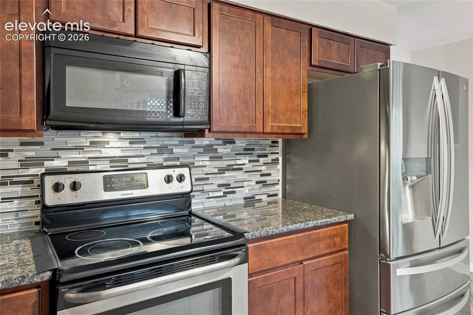 Modern kitchen featuring rich wood cabinetry, stainless steel appliances including a French-door refrigerator and electric range, granite countertops, and a geometric tile backsplash in neutral tones accented with contrasting stripes