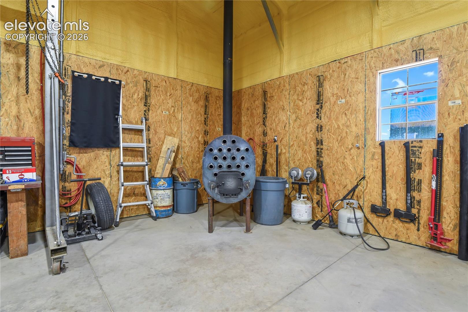 Workshop space featuring oriented strand board wall paneling, a concrete floor, a metal wood-burning stove, and a single-pane window
