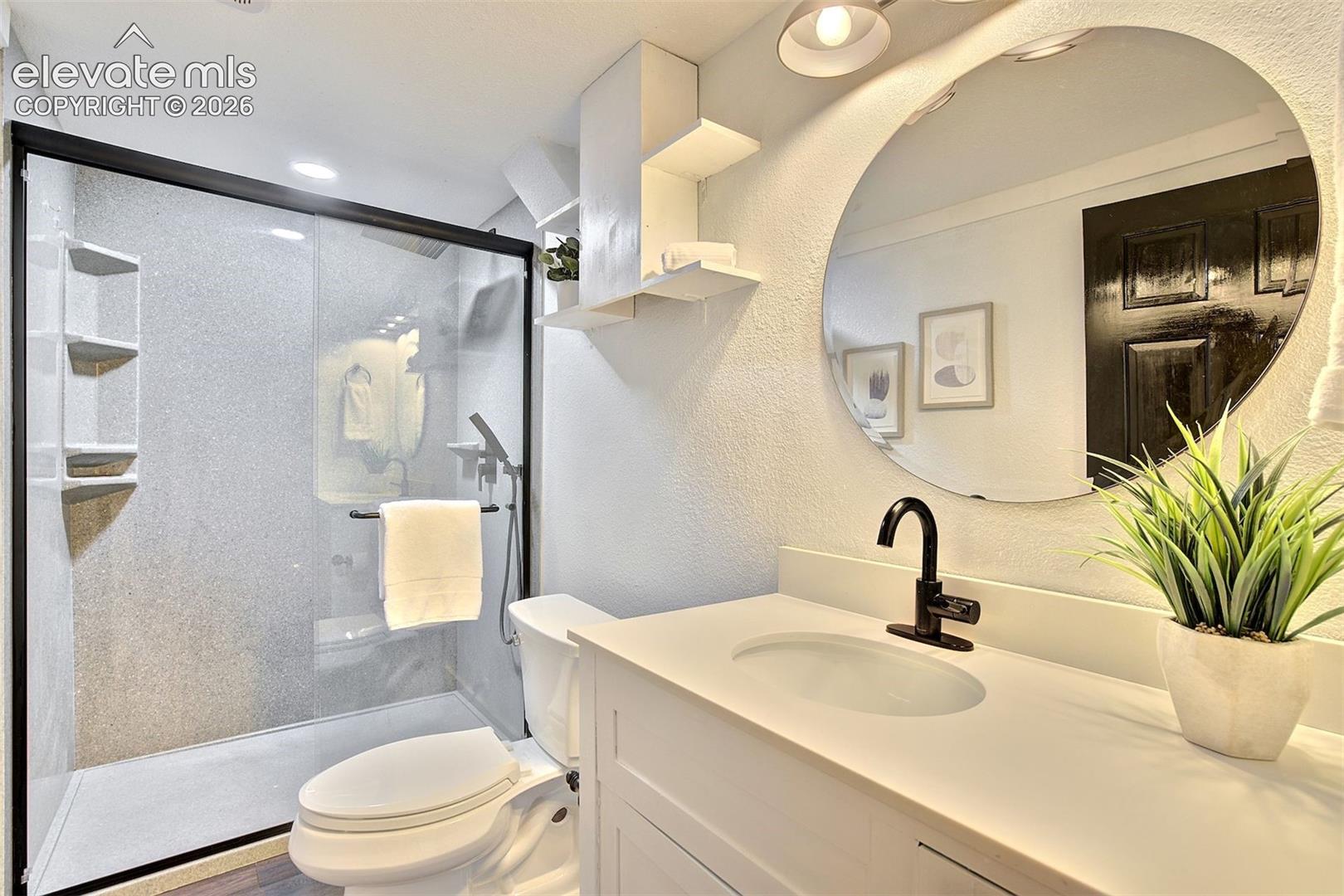 Bathroom featuring a walk-in shower with black-frame glass enclosure, custom wall-mounted shelving, solid-surface vanity countertop with under-mount sink, matte black faucet, and a circular wall mirror