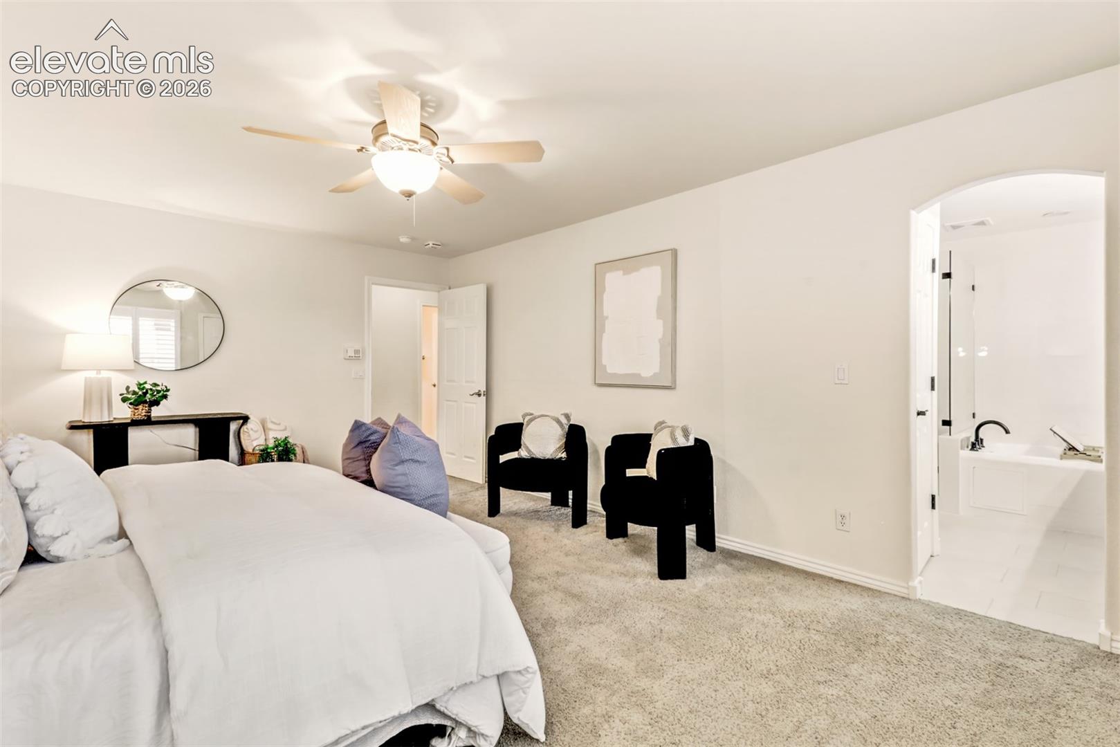 Neutral-toned wall-to-wall carpeting, ceiling fan with integrated lighting, and an arched doorway leading to a tiled bathroom featuring a soaking tub