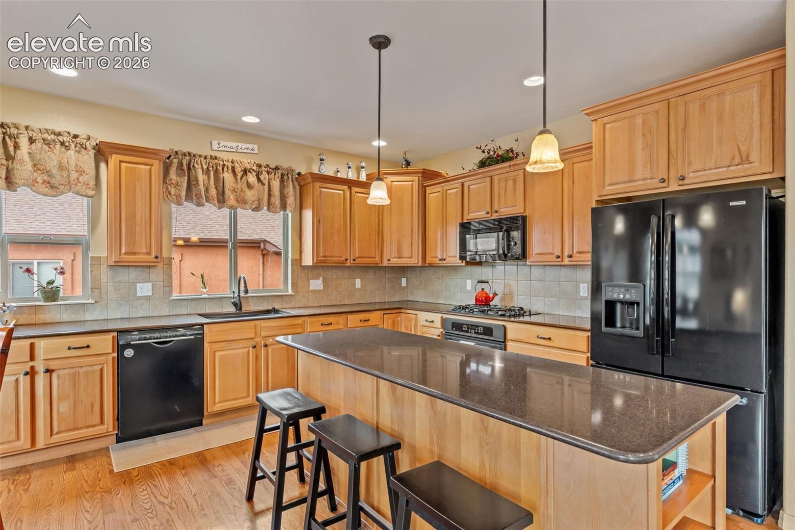 Kitchen featuring wood-finish flooring, light wood cabinetry, black appliances, a central island with a dark countertop, and recessed lighting