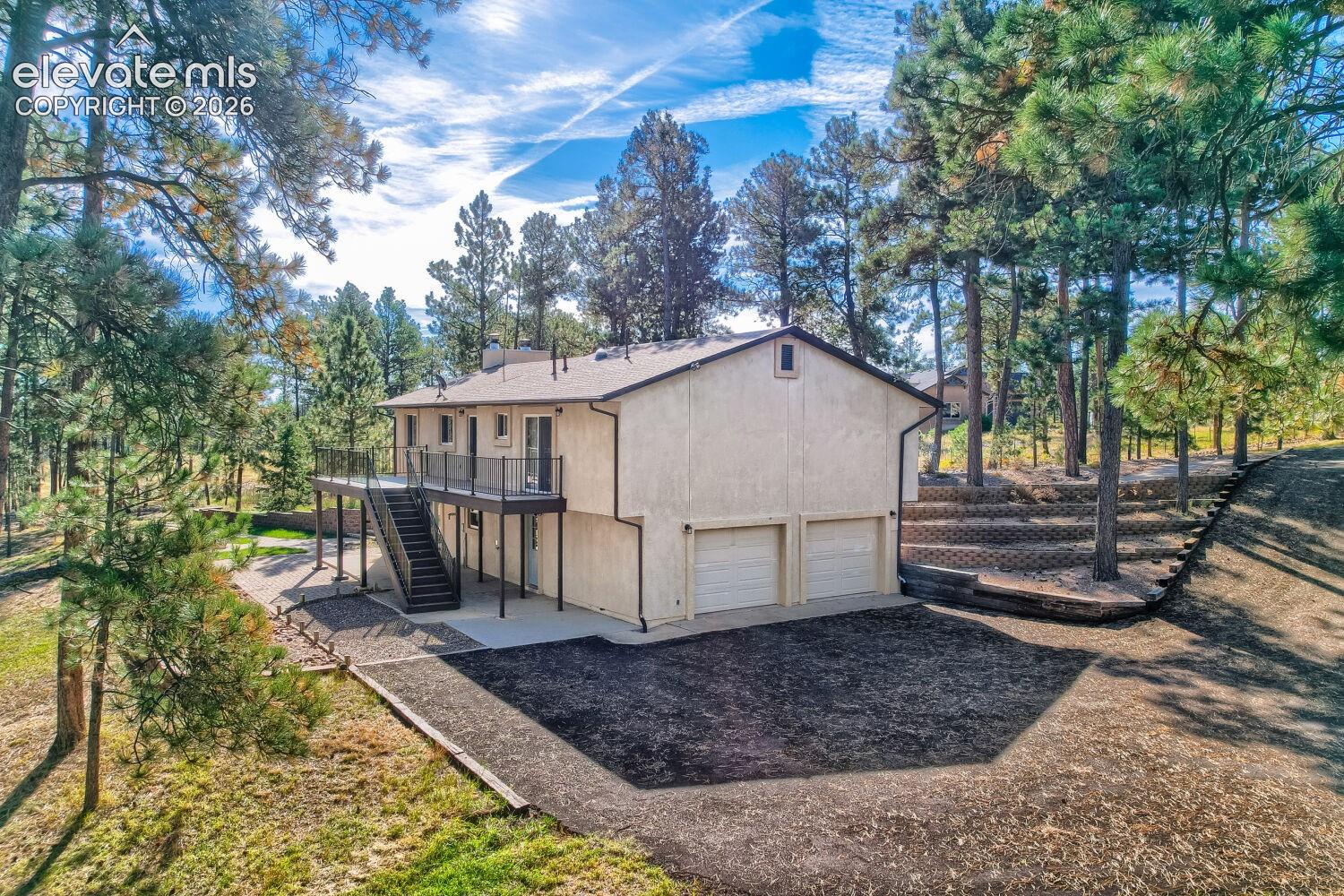 Two-car garage with white doors, expansive wood-finish deck with black railing, and terraced landscaping with mature trees