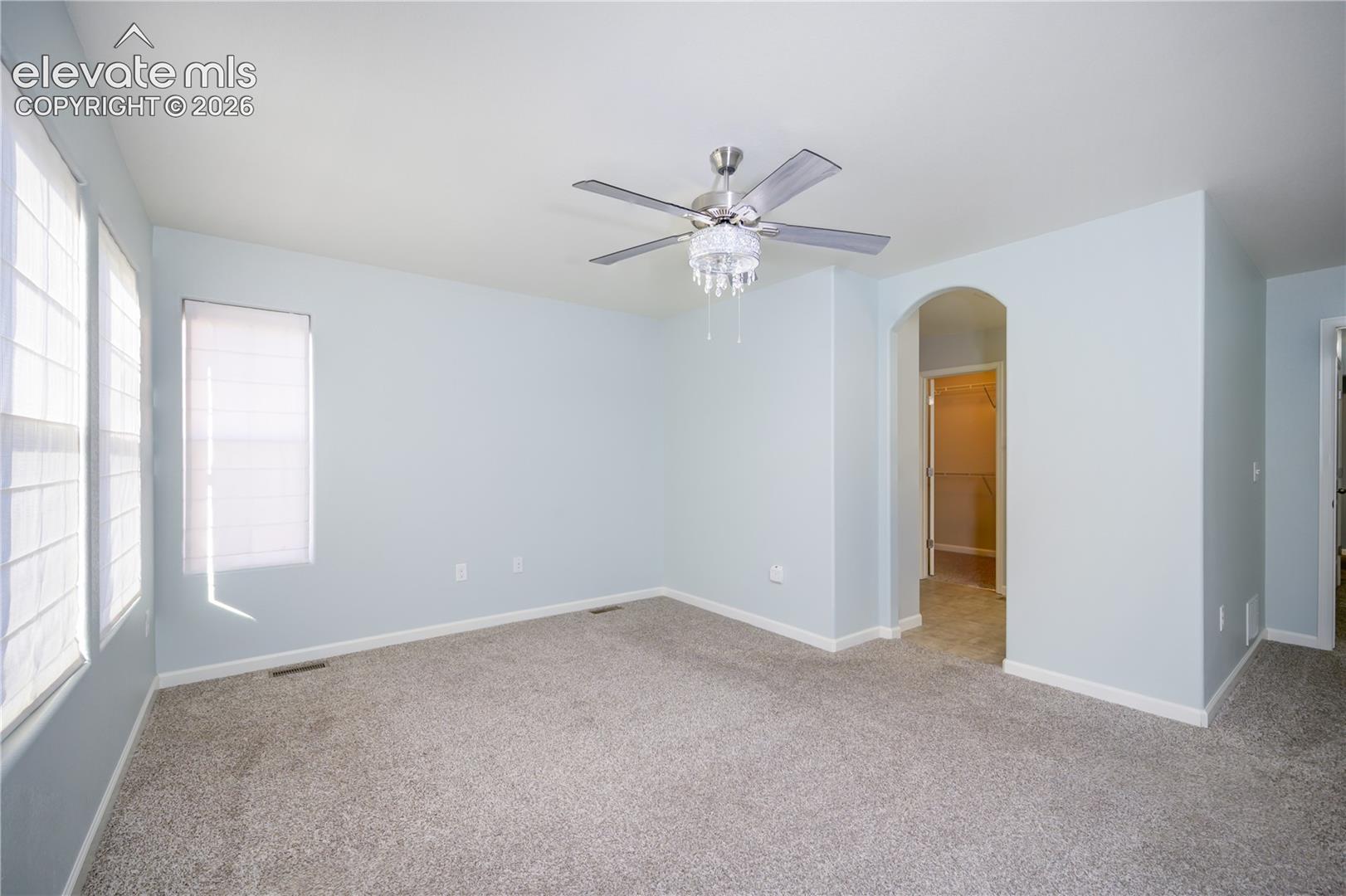 Neutral-toned wall-to-wall carpeting, ceiling fan with integrated crystal chandelier, multiple windows with blinds, and arched doorway entry