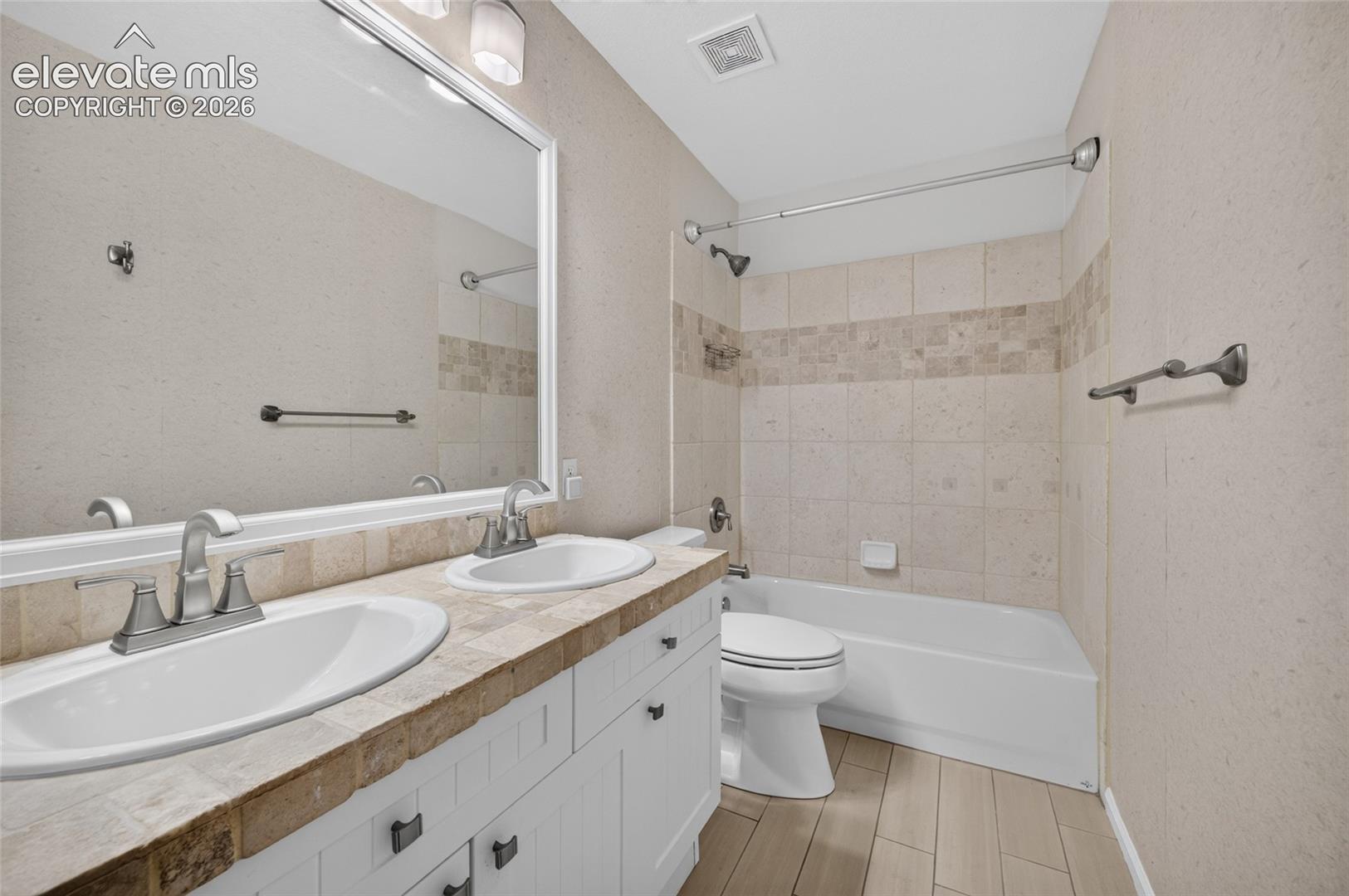 Bathroom featuring a dual-sink vanity with stone-tile countertops, wood-finish flooring, a large wall-mounted mirror, and a tiled shower-tub combination